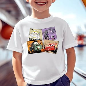 May include: White t-shirt featuring a colorful graphic of four cartoon cars in Halloween costumes. The cars are depicted as a mummy, a vampire, a zombie, and a car with flames. The design is set against a black background with Halloween-themed elements.