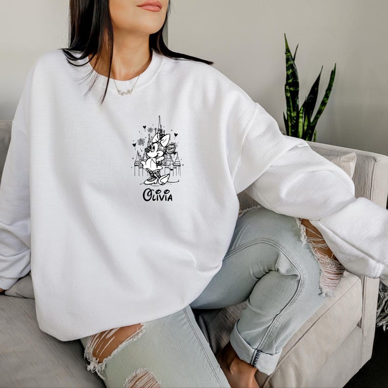 May include: White sweatshirt featuring a black and white graphic of a cartoon character in front of a city skyline. The name "Olivia" is written in cursive below the graphic. The sweatshirt has long sleeves and a crew neck.
