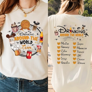 May include: White t-shirt with a graphic of a Mickey Mouse head with a spiderweb and the text "Drinking Around the World Team". The graphic includes images of drinks, snacks, and a Mickey Mouse head. The back of the shirt has the text "Drinking Around the World" with a list of countries and the word "Drunk".