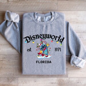 May include: A heather gray sweatshirt featuring a graphic of Stitch from Disney's Lilo & Stitch, wearing a Santa hat and holding Christmas lights. The sweatshirt has the text "Disneyworld" and "Florida" printed on it, with the year "1971".