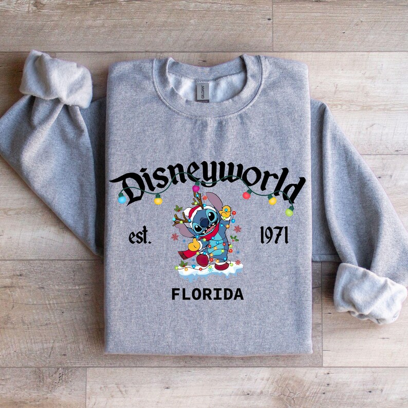 May include: A heather gray sweatshirt featuring a graphic of Stitch from Disney's Lilo & Stitch, wearing a Santa hat and holding Christmas lights. The sweatshirt has the text "Disneyworld" and "Florida" printed on it, with the year "1971".