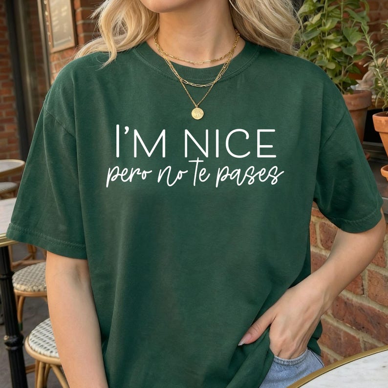 May include: A dark green short-sleeve t-shirt featuring the white text I'M NICE pero no Te pases printed on the front. The shirt has a crew neckline and a relaxed fit, showcasing a combination of bold block lettering and a cursive script font.