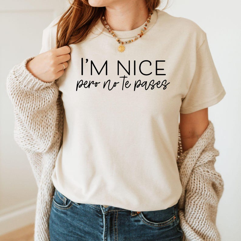 May include: A beige short-sleeved t-shirt featuring the black printed text I'M NICE pero no Te pases. The shirt has a crew neckline and is worn with a chunky knit cardigan and a beaded necklace with a small gold sun pendant.