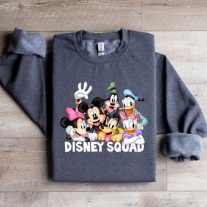 Puede incluir: Sudadera gris oscuro con un gr&aacute;fico colorido de personajes de Disney, incluyendo Mickey Mouse, Minnie Mouse, Donald Duck y Goofy. El texto "DISNEY SQUAD" est&aacute; impreso en blanco. La sudadera tiene cuello redondo y mangas largas.