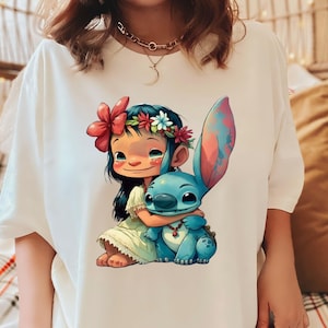 May include: White t-shirt with a colorful cartoon image of a girl with a flower crown hugging a blue alien character. The girl is wearing a green dress and the alien has a red necklace.