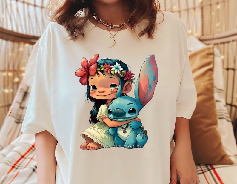 May include: White t-shirt with a colorful cartoon image of a girl with a flower crown hugging a blue alien character. The girl is wearing a green dress and the alien has a red necklace.