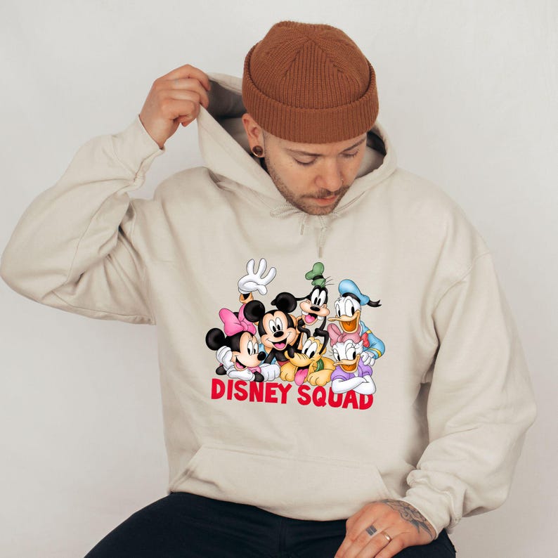 Puede incluir: Sudadera con capucha beige con un gr&aacute;fico colorido de personajes de Disney, incluyendo Mickey Mouse, Minnie Mouse, Donald Duck y Goofy. Las palabras "DISNEY SQUAD" est&aacute;n impresas en rojo. La sudadera tiene una capucha con cord&oacute;n.