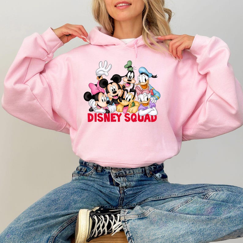 Puede incluir: Sudadera con capucha rosa claro con un estampado de personajes de Disney, incluyendo Mickey Mouse, Minnie Mouse, Donald Duck y Goofy. El texto "DISNEY SQUAD" est&aacute; impreso en rojo. La sudadera es usada por una persona.