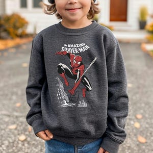 Puede incluir: Sudadera gris oscuro con un gr&aacute;fico de Spider-Man en acci&oacute;n, con el texto "the AMAZING SPIDER-MAN" arriba. La ilustraci&oacute;n muestra a Spider-Man balance&aacute;ndose de una telara&ntilde;a, con un paisaje urbano de fondo. Un art&iacute;culo de ropa para ni&ntilde;os.