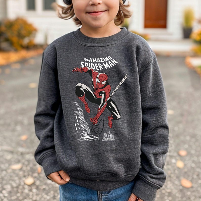 Puede incluir: Sudadera gris oscuro con un gr&aacute;fico de Spider-Man en acci&oacute;n, con el texto "the AMAZING SPIDER-MAN" arriba. La ilustraci&oacute;n muestra a Spider-Man balance&aacute;ndose de una telara&ntilde;a, con un paisaje urbano de fondo. Un art&iacute;culo de ropa para ni&ntilde;os.