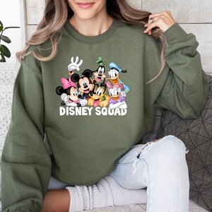 Puede incluir: Sudadera verde oliva con un gr&aacute;fico de personajes de Disney, incluyendo Mickey y Minnie Mouse, Goofy, Donald Duck y Pluto. Las palabras "DISNEY SQUAD" est&aacute;n impresas en blanco. La sudadera tiene cuello redondo.