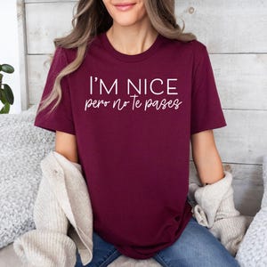 May include: A maroon short-sleeved t-shirt featuring the white text I'M NICE pero no te pases printed on the front. The shirt has a crew neckline and a standard fit, paired with a beige textured cardigan draped over the arms.