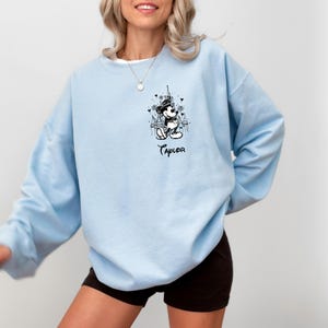 May include: Light blue sweatshirt featuring a black and white graphic of Mickey Mouse in front of a castle, with the name "Taylor" below. The sweatshirt has long sleeves and a crew neck.