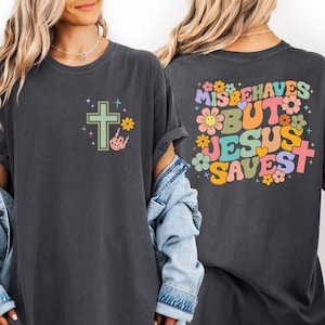 May include: Dark gray t-shirt with a colorful graphic on the front and back. The front features a cross, flower, and skeleton hand. The back says "Misbehaves But Jesus Saves" with flowers and crosses.