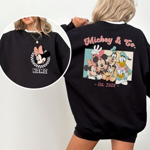 Op de afbeelding: Zwart sweatshirt met Minnie Mouse, roze strik en een geblokte cirkel, met de tekst "NAME" eronder. De achterkant toont Mickey & Co. personages en de tekst "Est. 1928". Het sweatshirt heeft een witte kraag.