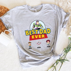 K&ouml;nnte beinhalten: Hellgraues T-Shirt mit dem Schriftzug "BEST DAD EVER" in Gelb und Rot, mit einem Cartoon-Bild von Buzz Lightyear. Darunter die Zeichentrickfiguren Alex, Jessi und Mary.