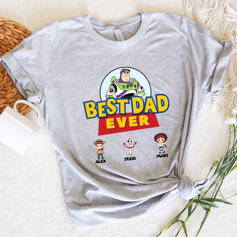 K&ouml;nnte beinhalten: Hellgraues T-Shirt mit dem Schriftzug "BEST DAD EVER" in Gelb und Rot, mit einem Cartoon-Bild von Buzz Lightyear. Darunter die Zeichentrickfiguren Alex, Jessi und Mary.