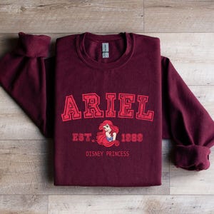 May include: A maroon sweatshirt featuring the name "ARIEL" in red, block lettering. Below the name is a graphic of Ariel, the Disney princess, with the text "EST. 1989 DISNEY PRINCESS". The sweatshirt is folded on a wooden surface.
