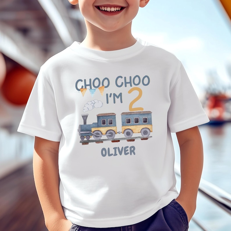 May include: A white short-sleeved t-shirt featuring a watercolor-style graphic of a blue and yellow train. The text reads CHOO CHOO I'M 2 with the name OLIVER printed below the train. Small colorful pennant flags and a cloud of smoke are included.