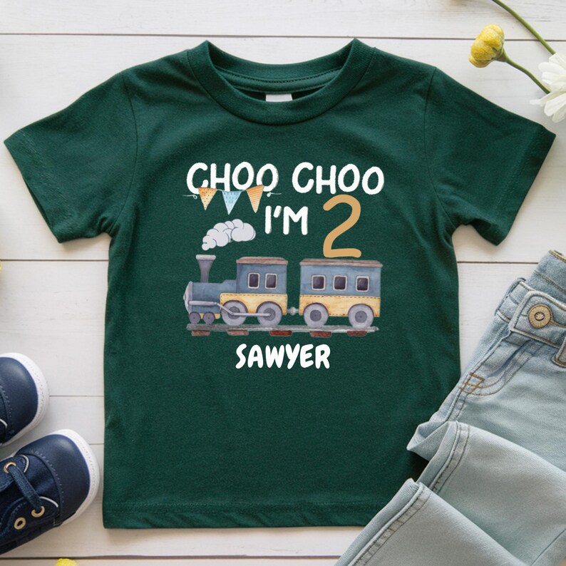 May include: A dark green short-sleeve t-shirt featuring a graphic of a train with the text "CHOO CHOO I'M 2" and the name "SAWYER" printed in white. The design includes a large tan number two and small colorful pennant flags above the train.