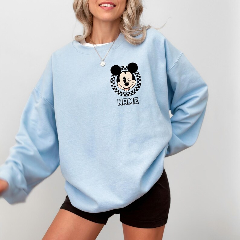 Op de afbeelding: Lichtblauw sweatshirt met een Mickey Mouse-ontwerp, geplaatst in een geblokte cirkel. Het woord "NAME" staat eronder gedrukt. De sweatshirt heeft een ronde hals en lange mouwen.