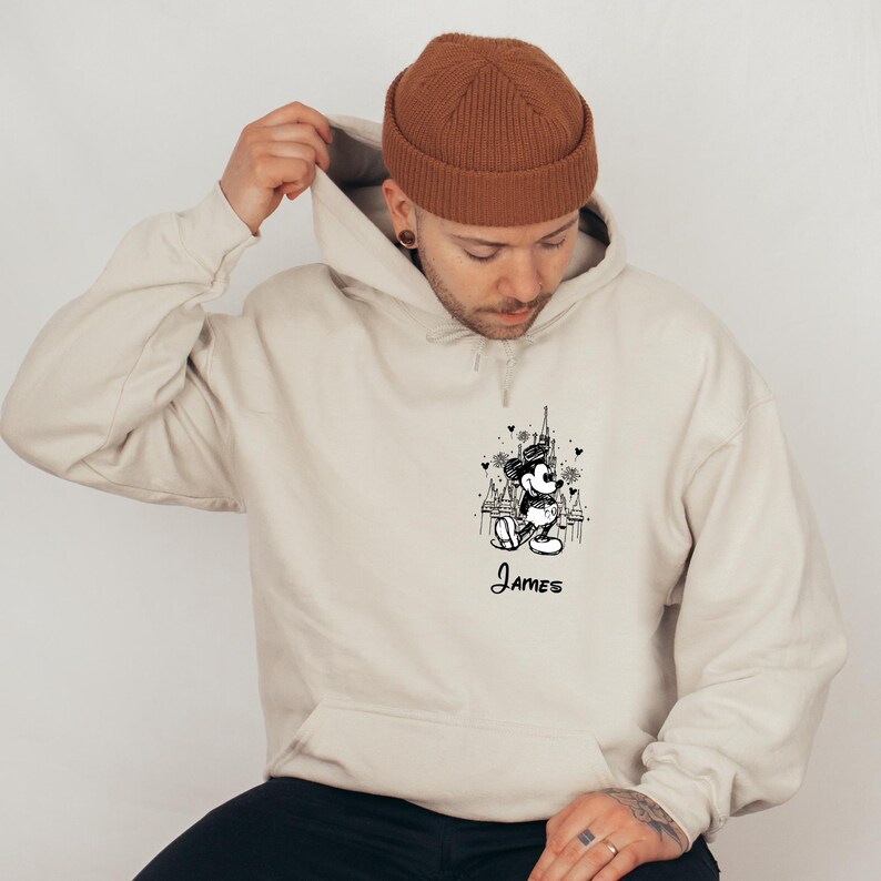 May include: A beige hoodie featuring a black and white illustration of a cartoon character with the name "James" below. The hoodie has a drawstring hood and a kangaroo pocket.