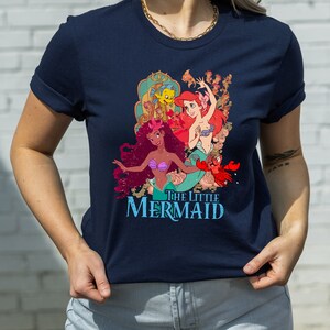 May include: A navy blue t-shirt featuring a vintage-style graphic of Ariel and her sisters from The Little Mermaid. The graphic depicts the characters in a colourful, whimsical style.