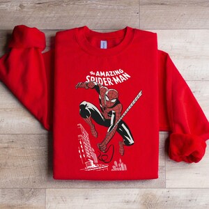 Puede incluir: Sudadera roja con un dise&ntilde;o de Spider-Man en acci&oacute;n, con el texto "The Amazing Spider-Man" en blanco. El dise&ntilde;o incluye un paisaje urbano. La sudadera tiene cuello redondo y mangas largas.