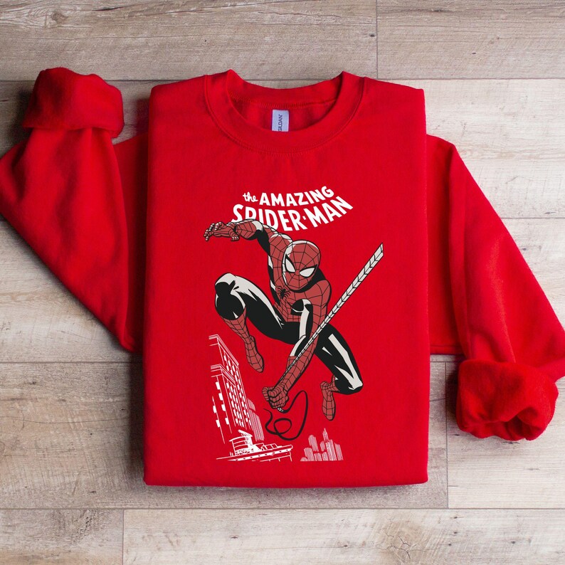 Puede incluir: Sudadera roja con un dise&ntilde;o de Spider-Man en acci&oacute;n, con el texto "The Amazing Spider-Man" en blanco. El dise&ntilde;o incluye un paisaje urbano. La sudadera tiene cuello redondo y mangas largas.
