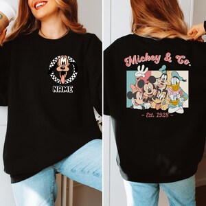 Op de afbeelding: Zwart sweatshirt met een afbeelding van Pluto in een geruite cirkel en het woord "NAME" eronder. De achterkant van de trui toont "Mickey & Co." met Disney-figuren en de tekst "- Est. 1928 -."