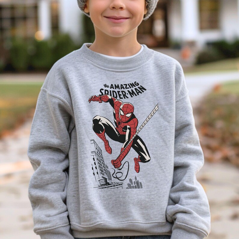 Puede incluir: Una sudadera gris claro con un gr&aacute;fico de Spider-Man en una pose din&aacute;mica. El dise&ntilde;o incluye el texto "The Amazing Spider-Man" en blanco. El superh&eacute;roe est&aacute; representado con su cl&aacute;sico traje rojo y negro, con una telara&ntilde;a que se extiende desde su mano.