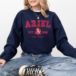 May include: A navy blue sweatshirt with the name "ARIEL" printed in red block letters. Underneath, it reads "EST. 1989" with a graphic of Ariel, the Disney Princess. The words "DISNEY PRINCESS" are printed below the graphic.