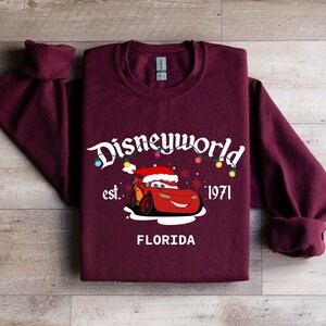 May include: Burgundy sweatshirt featuring a graphic of Lightning McQueen wearing a Santa hat, with the text "Disneyworld est. 1971 FLORIDA". The design includes colorful Christmas lights and snowflakes.
