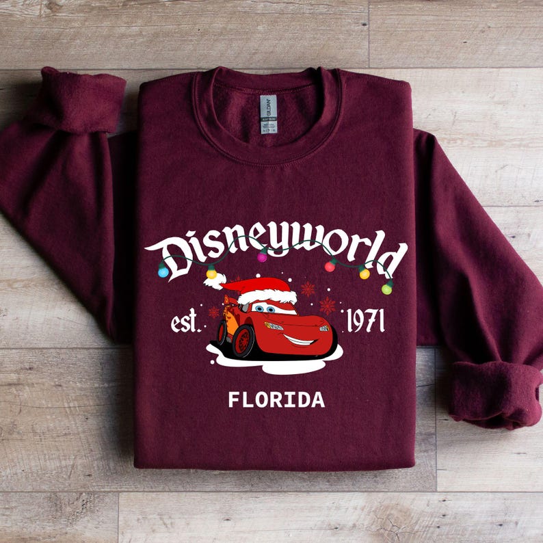 May include: Burgundy sweatshirt featuring a graphic of Lightning McQueen wearing a Santa hat, with the text "Disneyworld est. 1971 FLORIDA". The design includes colorful Christmas lights and snowflakes.