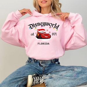 May include: A light pink hooded sweatshirt featuring the text "Disneyworld" with a cartoon car wearing a Santa hat. The sweatshirt also includes the text "est. 1971" and "FLORIDA".