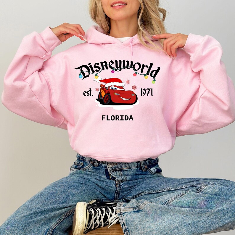 May include: A light pink hooded sweatshirt featuring the text "Disneyworld" with a cartoon car wearing a Santa hat. The sweatshirt also includes the text "est. 1971" and "FLORIDA".