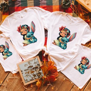 May include: Four white t-shirts with a cartoon image of a girl holding a blue alien character. The girl is wearing a flower crown and a white dress. The alien character has large ears and is holding a heart.