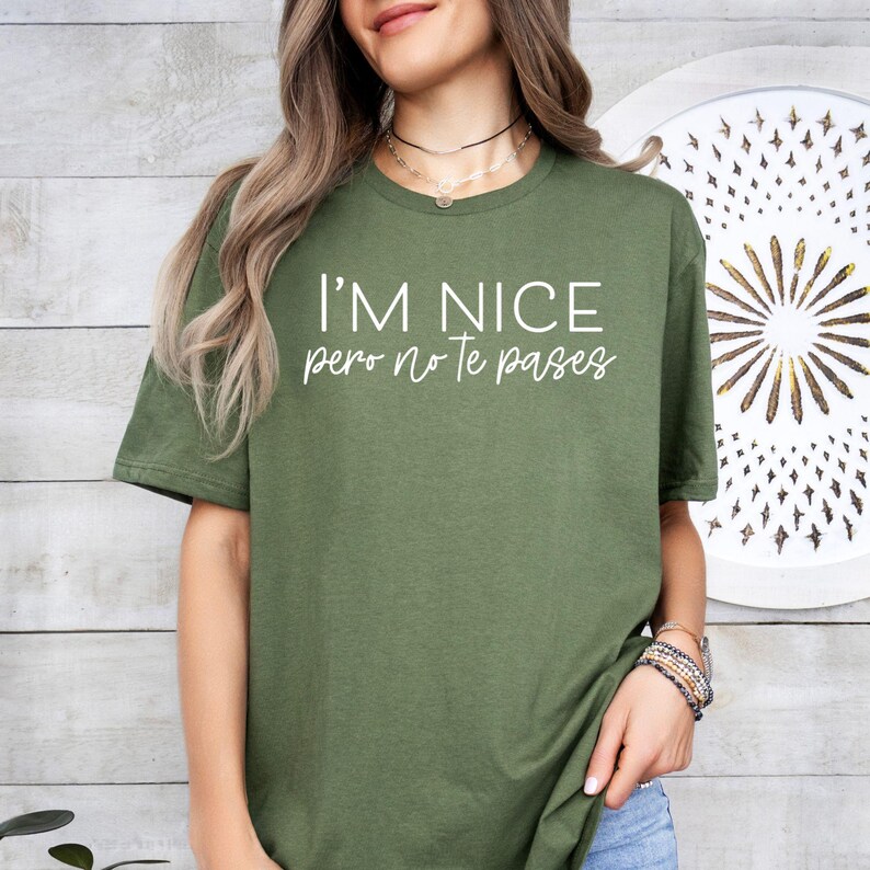 May include: An olive green t-shirt featuring the white text I'M NICE pero no Te pases printed on the front. The shirt has short sleeves and a crew neckline, providing a simple and straightforward design for casual wear in a standard fit.