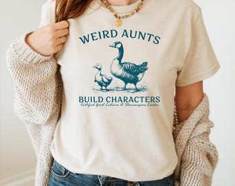 May include: A beige short-sleeve t-shirt featuring a dark teal graphic of a goose and a gosling. The text reads WEIRD AUNTS BUILD CHARACTERS Certified Great Listener & Shenanigans Leader in a dark teal font centered above and below the illustration.