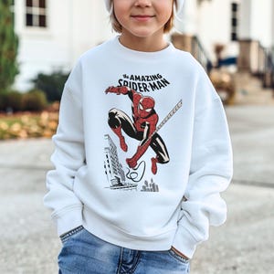 Puede incluir: Sudadera blanca con un dise&ntilde;o de Spider-Man en acci&oacute;n. El superh&eacute;roe, con su traje rojo y negro, se balancea en una telara&ntilde;a. El texto "The Amazing Spider-Man" est&aacute; sobre la imagen.