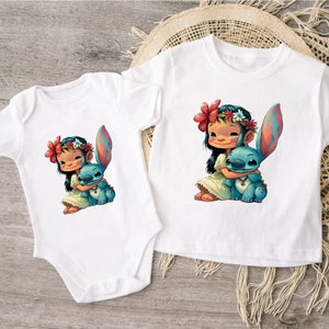 May include: Two white t-shirts with a cartoon image of a girl with long black hair wearing a flower crown and holding a blue alien character. The girl is wearing a white dress and the alien is blue with red accents.