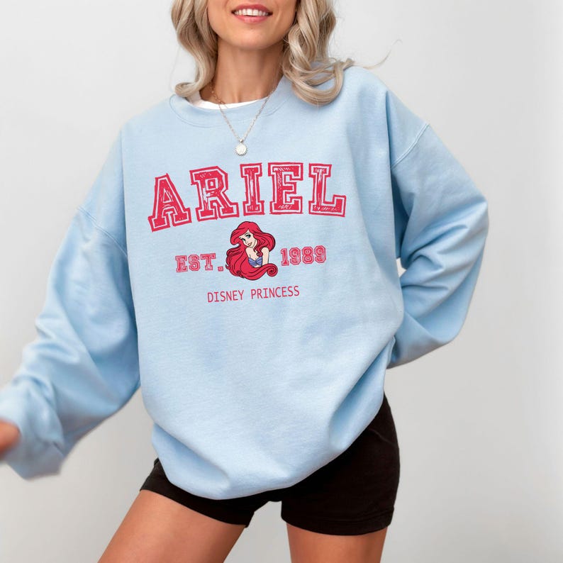 May include: A pale blue sweatshirt with the name "ARIEL" in red lettering. Below the name is a graphic of Ariel, the Disney princess, with the text "EST. 1989 DISNEY PRINCESS". The sweatshirt has a crew neck and long sleeves.