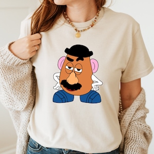 May include: A beige t-shirt with a cartoon image of Mr. Potato Head with a black hat, brown eyes, a brown nose, a black mustache, and blue pants.