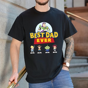 K&ouml;nnte beinhalten: Schwarzes T-Shirt mit dem Schriftzug "BEST DAD EVER" in Gelb und Blau, auf einem roten Banner. Das Design zeigt Zeichentrickfiguren und Buzz Lightyear. Darunter befinden sich vier Zeichentrickfiguren mit dem Wort "NAME" darunter.