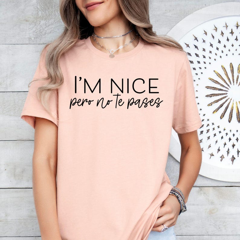 May include: A light peach-colored short-sleeve t-shirt featuring the black printed text I'M NICE pero no te pases centered on the chest. The garment has a classic crew neckline and a standard fit, providing a simple and clean appearance.