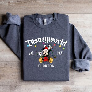 May include: A dark gray sweatshirt featuring the text "Disneyworld" above a graphic of Mickey Mouse wearing a Santa hat and tangled in Christmas lights. The text "est. 1971 FLORIDA" is below the graphic.