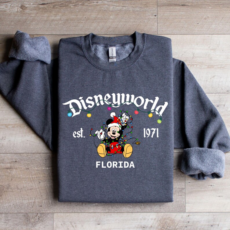 May include: A dark gray sweatshirt featuring the text "Disneyworld" above a graphic of Mickey Mouse wearing a Santa hat and tangled in Christmas lights. The text "est. 1971 FLORIDA" is below the graphic.
