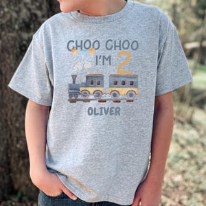 May include: A heather gray short-sleeve t-shirt featuring a graphic of a blue and yellow train on tracks. The text reads CHOO CHOO I'M 2 OLIVER, accompanied by small colorful pennant flags and a cloud illustration printed on the front of the shirt.