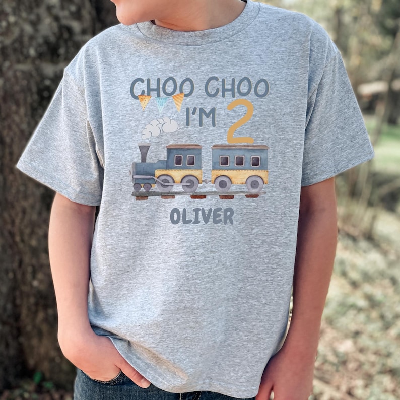 May include: A heather gray short-sleeve t-shirt featuring a graphic of a blue and yellow train on tracks. The text reads CHOO CHOO I'M 2 OLIVER, accompanied by small colorful pennant flags and a cloud illustration printed on the front of the shirt.