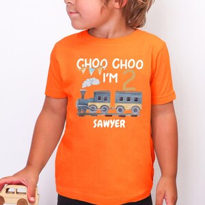 May include: A bright orange short-sleeved t-shirt featuring a graphic of a blue and yellow train with a puff of smoke. The text reads CHOO CHOO I'M 2 SAWYER in white letters, accompanied by a small string of colorful triangular bunting flags.